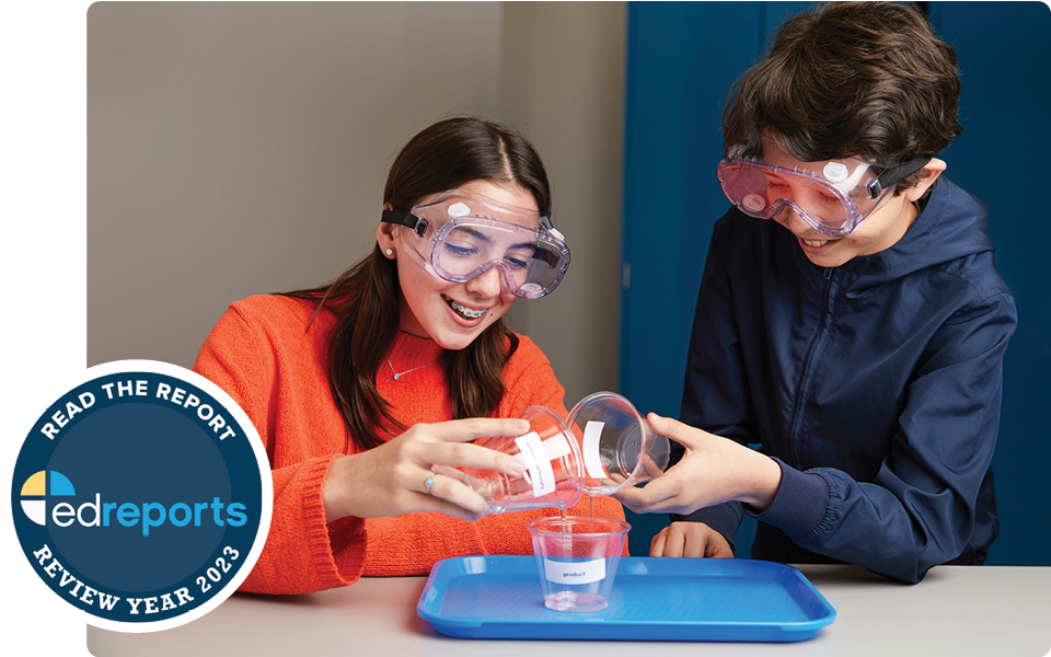 Two students wearing safety goggles conduct a science experiment together, pouring liquid between plastic cups over a blue tray on a classroom table.