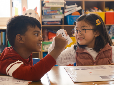 A boy and a girl sit at a classroom table, smiling as the girl holds up a flashcard with the letter "a" on it. Shelves with books and toys are visible in the background.