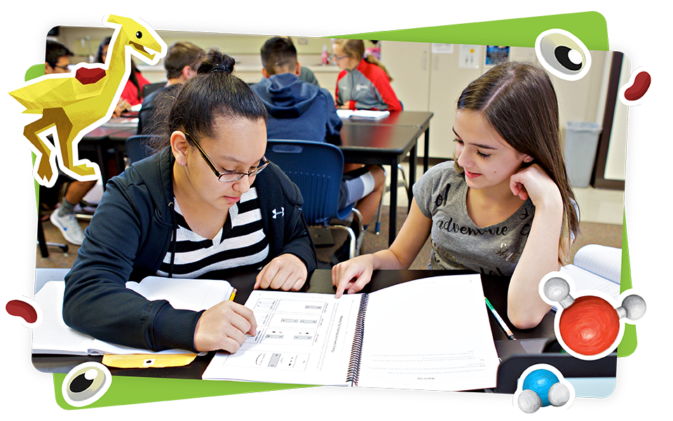 Two students sit at a classroom table working together on an assignment, surrounded by papers and books, with colorful cartoon graphics decorating the photo.