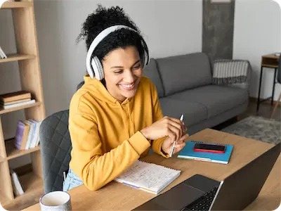 A woman wearing headphones and a yellow hoodie sits at a desk with a notebook, pen, mug, and laptop, smiling as she participates in a Virtual Tutoring Program for U.S. Tutors during an online meeting or virtual class.