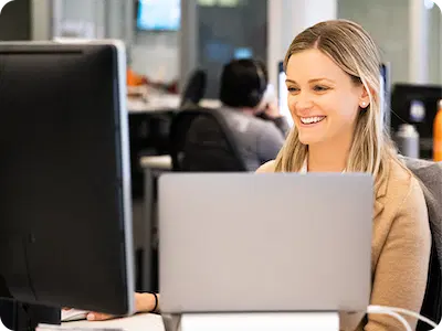 A woman sits at a desk in an office, smiling at a computer monitor with her laptop open, participating in a Virtual Tutoring Program for U.S. Tutors. Other people are visible working in the background.