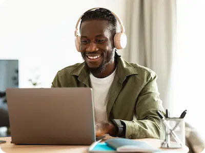 Man wearing headphones sits at a desk, smiling and working on a laptop for a Virtual Tutoring Program for U.S. Tutors, with books and a pen holder nearby.