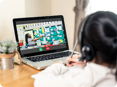 A student wearing headphones attends a Virtual Tutoring Program for U.S. Tutors, watching a teacher point at a green bulletin board on a laptop screen.