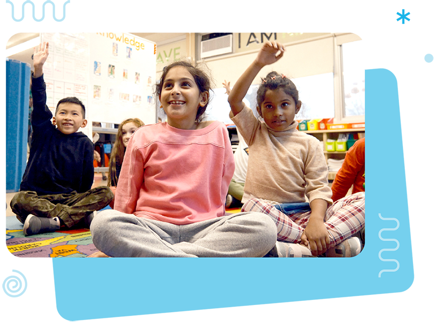 Four young children sit on a classroom floor, two with their hands raised, participating in an activity. Shelves of books and educational posters highlight the classroom’s focus on bilingual programs in the background.