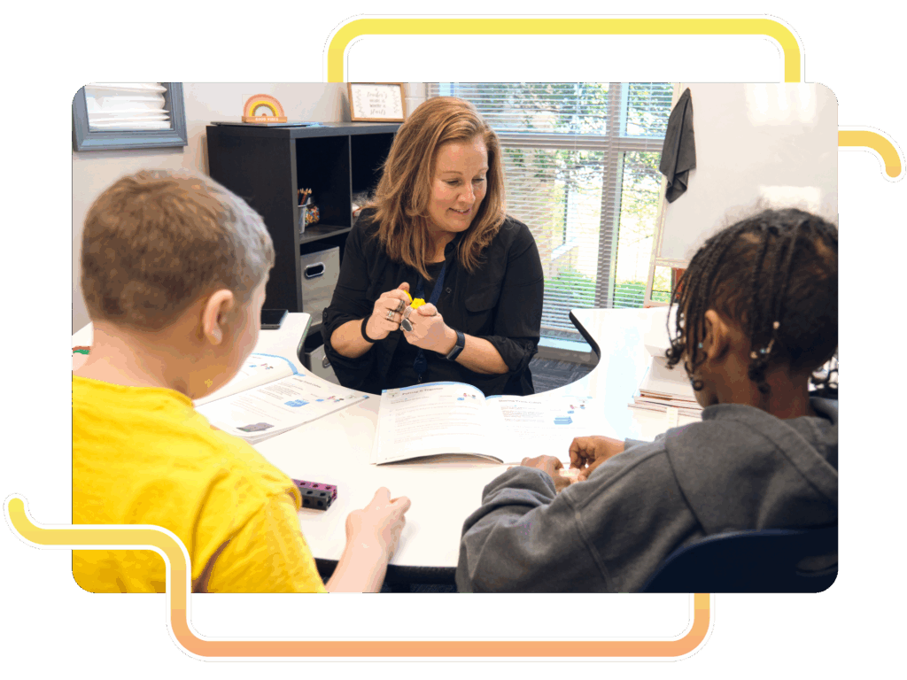 A teacher sits at a table with two students, using small objects and a workbook to provide individualized instruction during a Boost Math lesson in a classroom with large windows.