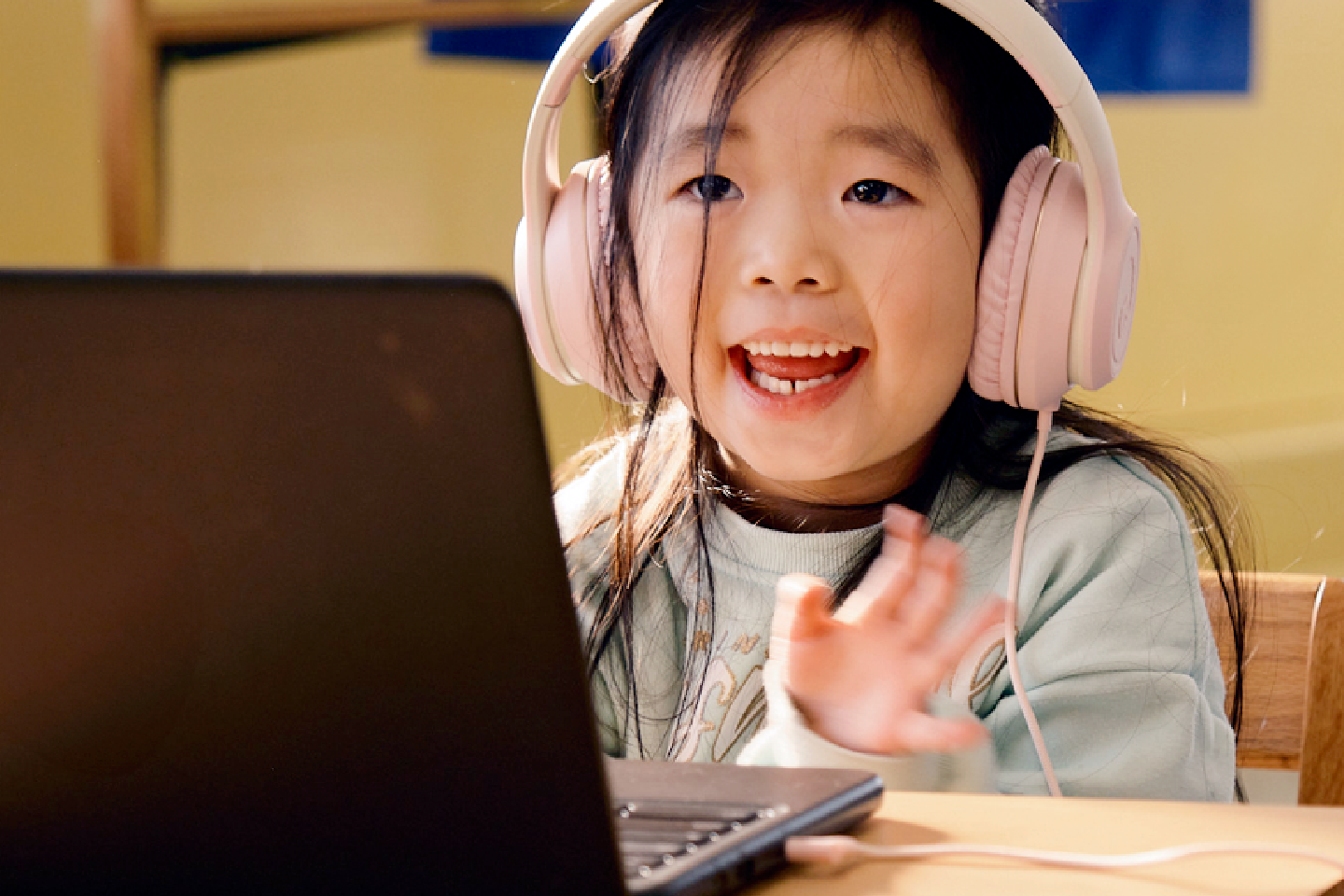 Young girl wearing headphones sits at a table, smiling and waving while looking at a laptop screen, ready to ampliar el plan de estudios through interactive online learning.