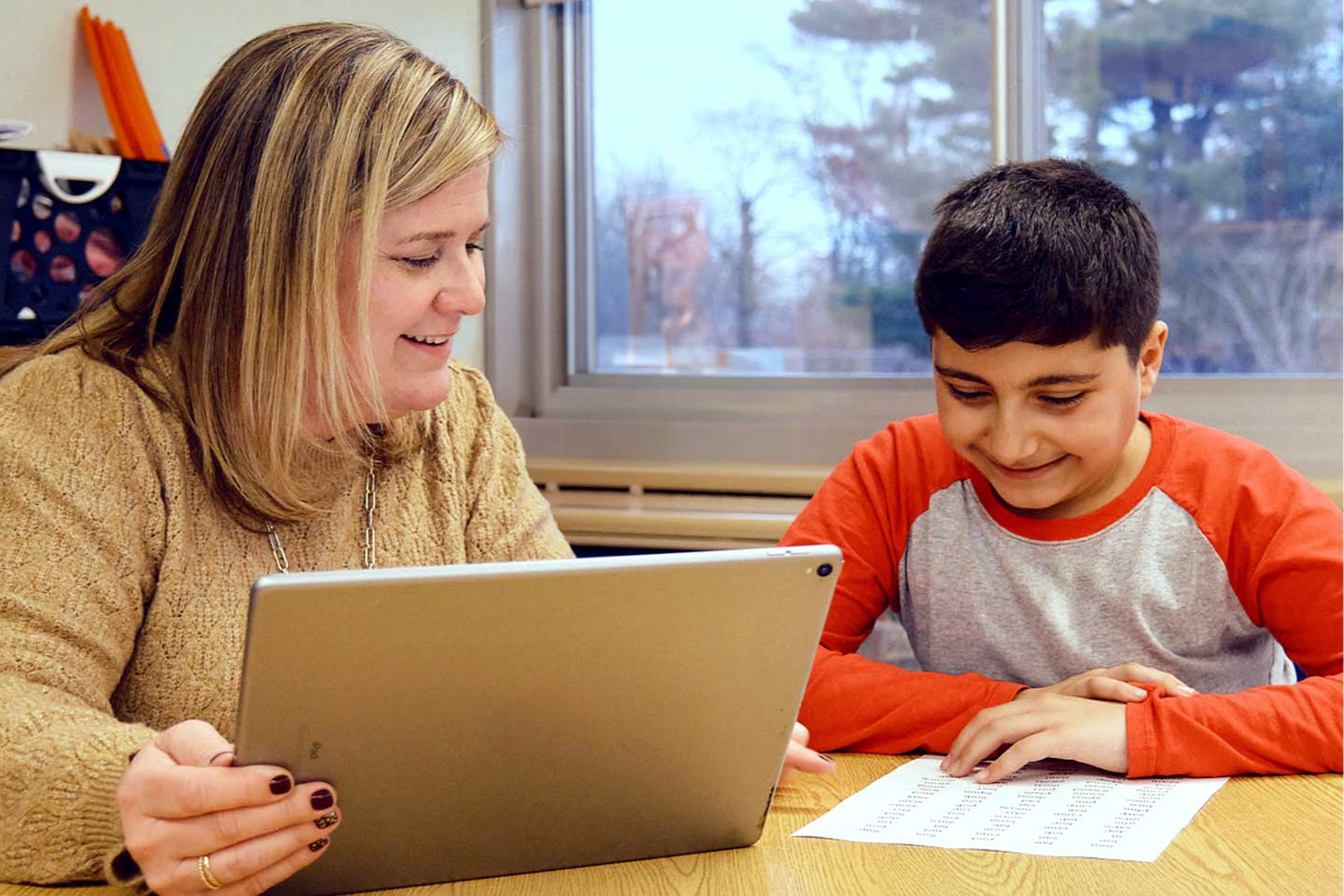 A woman shows a tablet to a boy sitting at a desk; the boy is smiling and looking at a sheet of printed text, exploring nuevas formas de ampliar el plan de estudios.