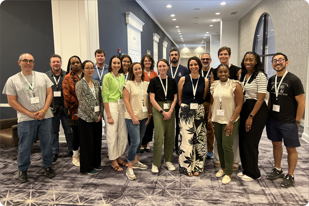 A collaborative community of sixteen people, including a Math teacher, pose together indoors on a carpeted hallway floor with neutral walls, all wearing conference badges.