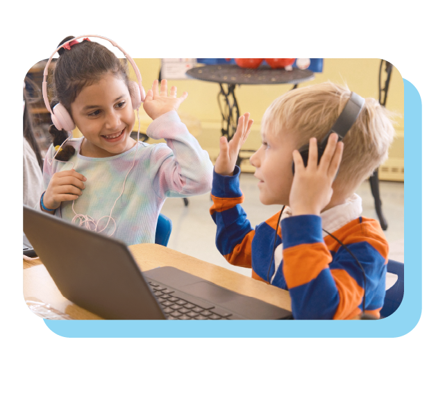 Two young children wearing headphones sit at a table with a laptop, smiling and giving each other a high five.