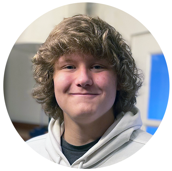A young person with wavy, light brown hair and a white hoodie smiles at the camera indoors.