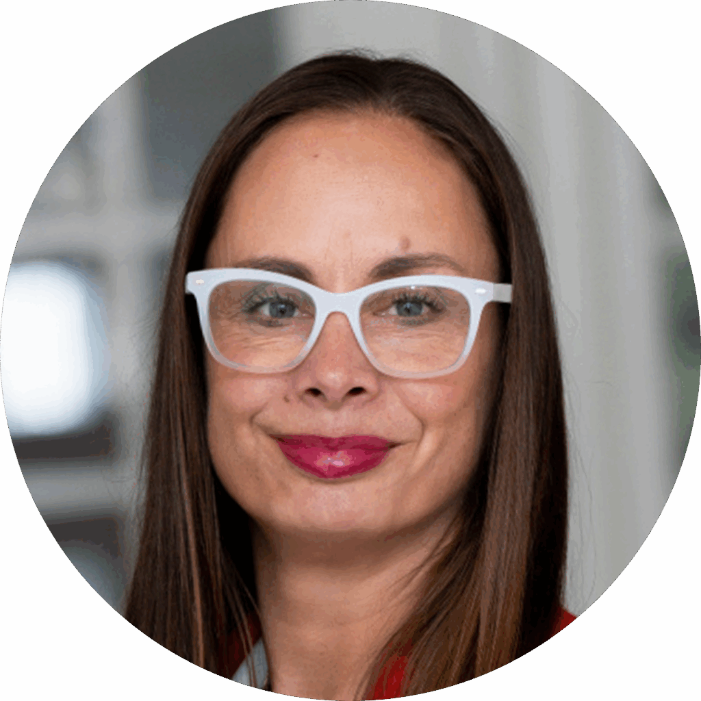 A woman with long brown hair, wearing white-framed glasses and a red top, is smiling at the camera against a blurred indoor background.