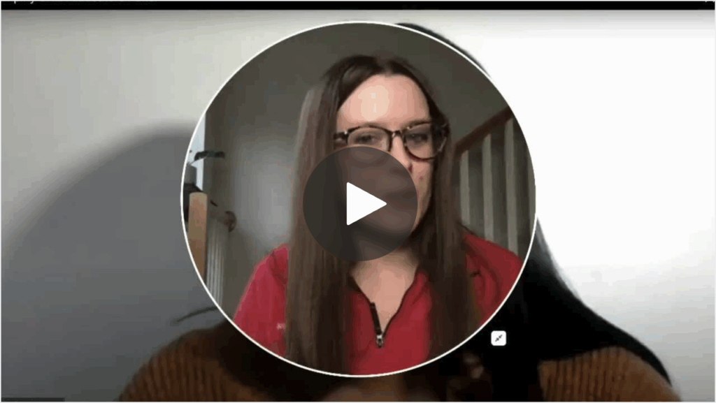 Screenshot of a paused video call showing a woman with long brown hair and glasses, wearing a red top, with a play button overlay in the center.