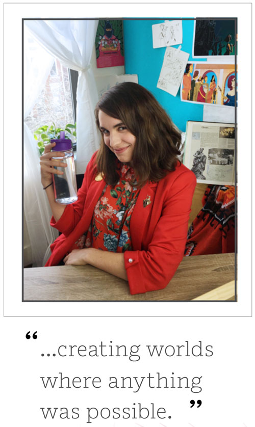 Young woman in a red blazer smiling at camera, holding a purple tumbler, seated at a desk with sketches on the wall behind her.