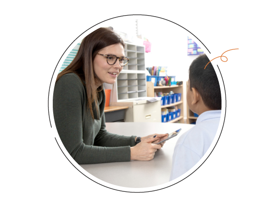 A teacher and a young student engage in conversation at a desk, with the teacher holding a tablet and pointing towards it.