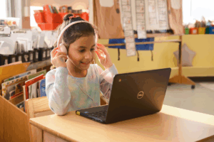 A young girl sits at a desk wearing headphones and using a laptop in a brightly lit classroom, focusing on her mCLASS Math assessment to support math progress monitoring.