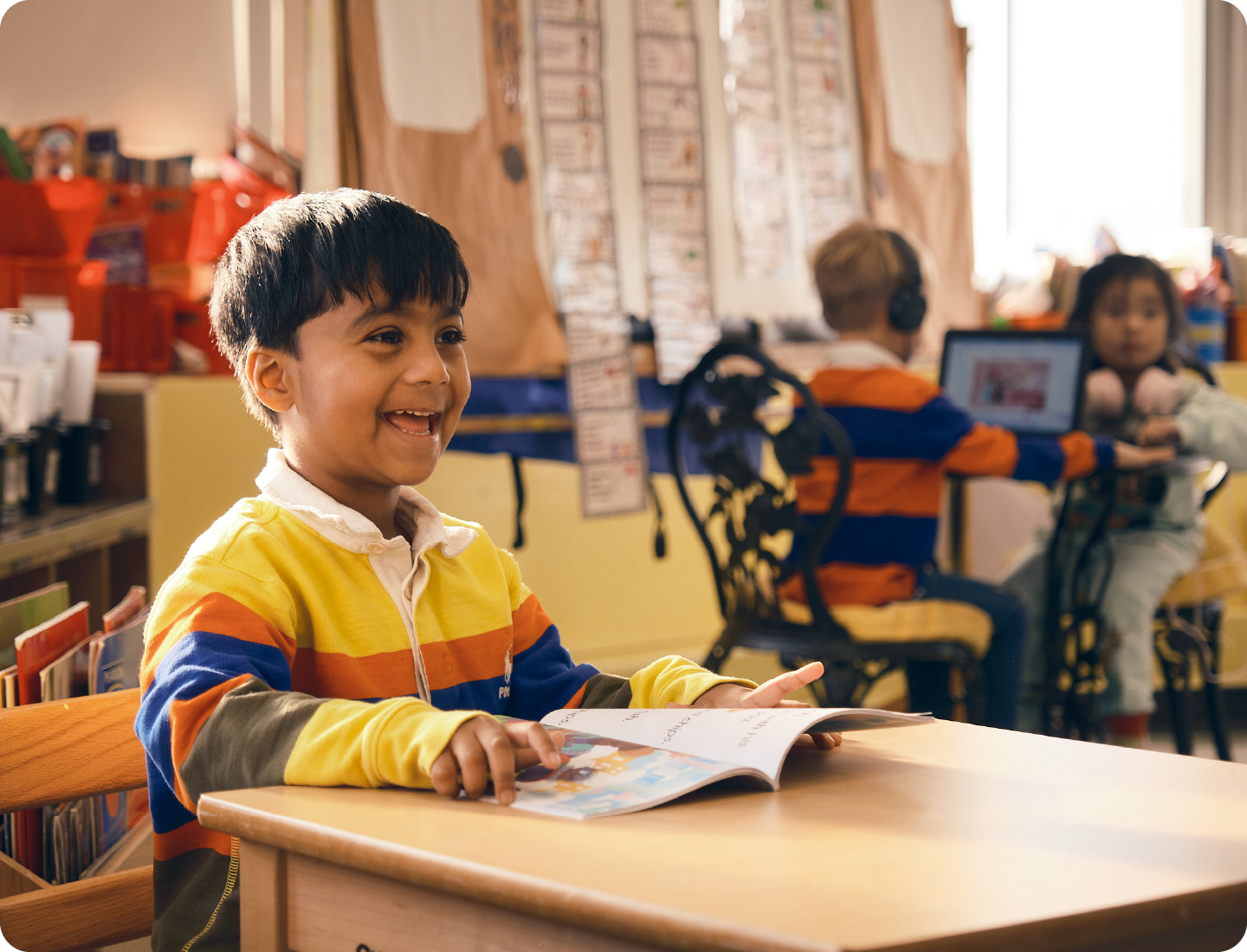 Smiling young boy sits at a classroom desk holding an open book, developing reading comprehension, while two other children are seen in the background engaged in activities.