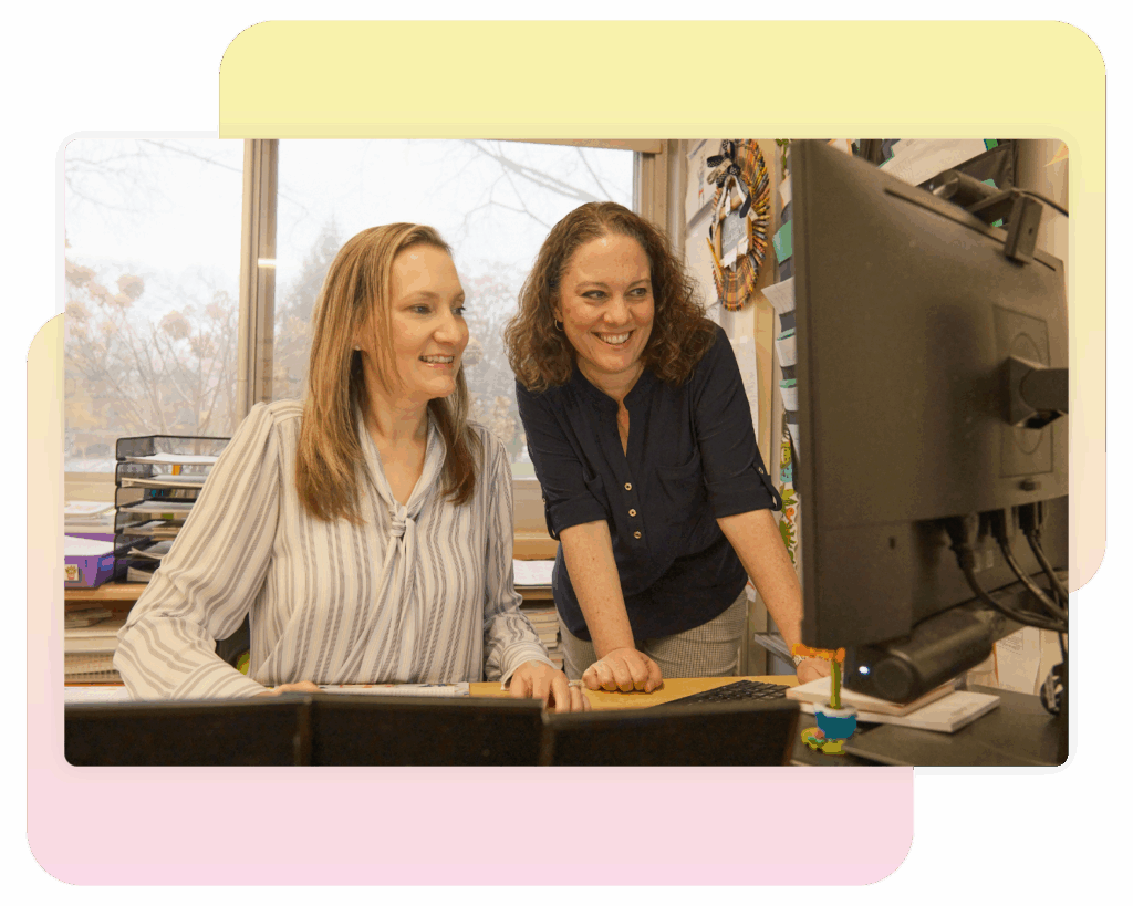 Two women in an office setting look at a computer monitor together, both appearing focused and engaged. One is seated while the other stands beside her.