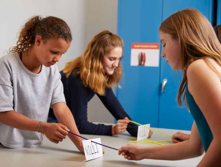 Three girls stand at a table in a classroom, using colored rods and cards labeled "VILLI" for a hands-on science activity.