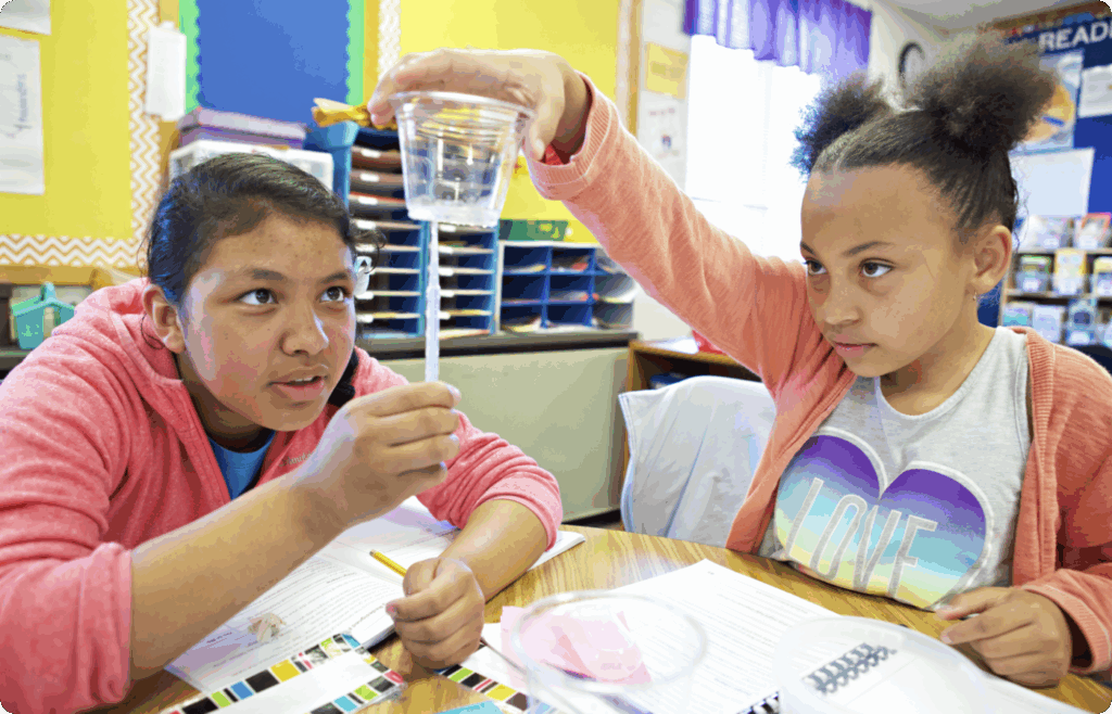 Two girls conduct a science experiment in a classroom, holding a clear cup with water and a straw, while observing and taking notes at their desks.