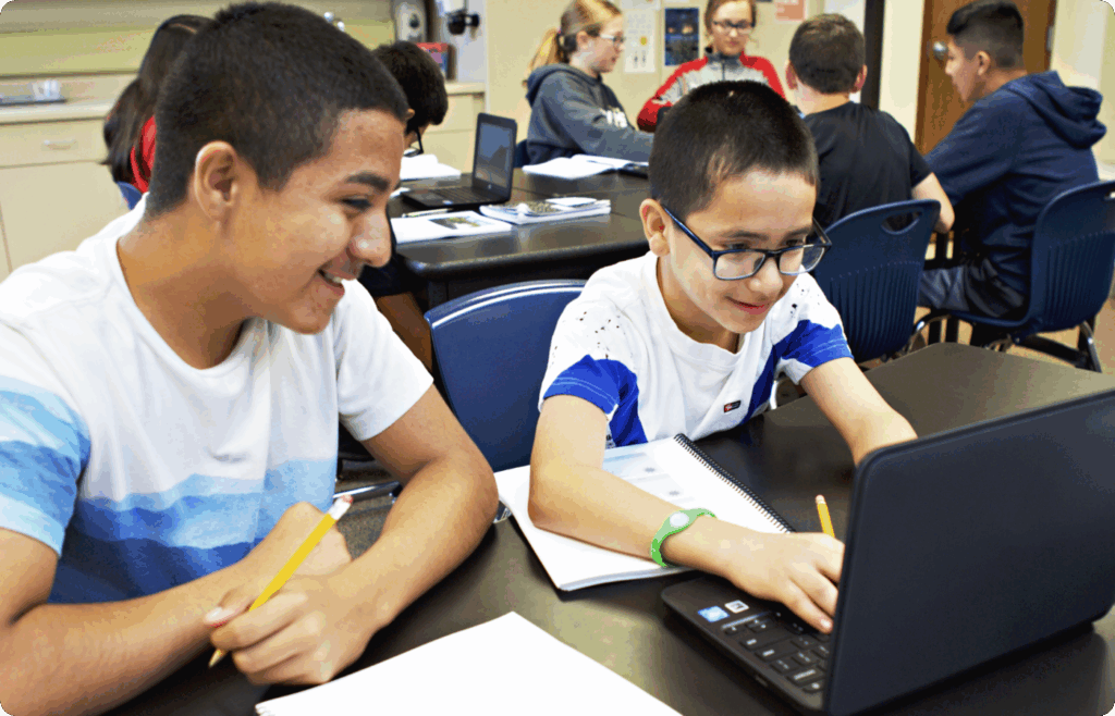 Two boys work together at a desk with a laptop and notebooks, while other students sit and work in the background.