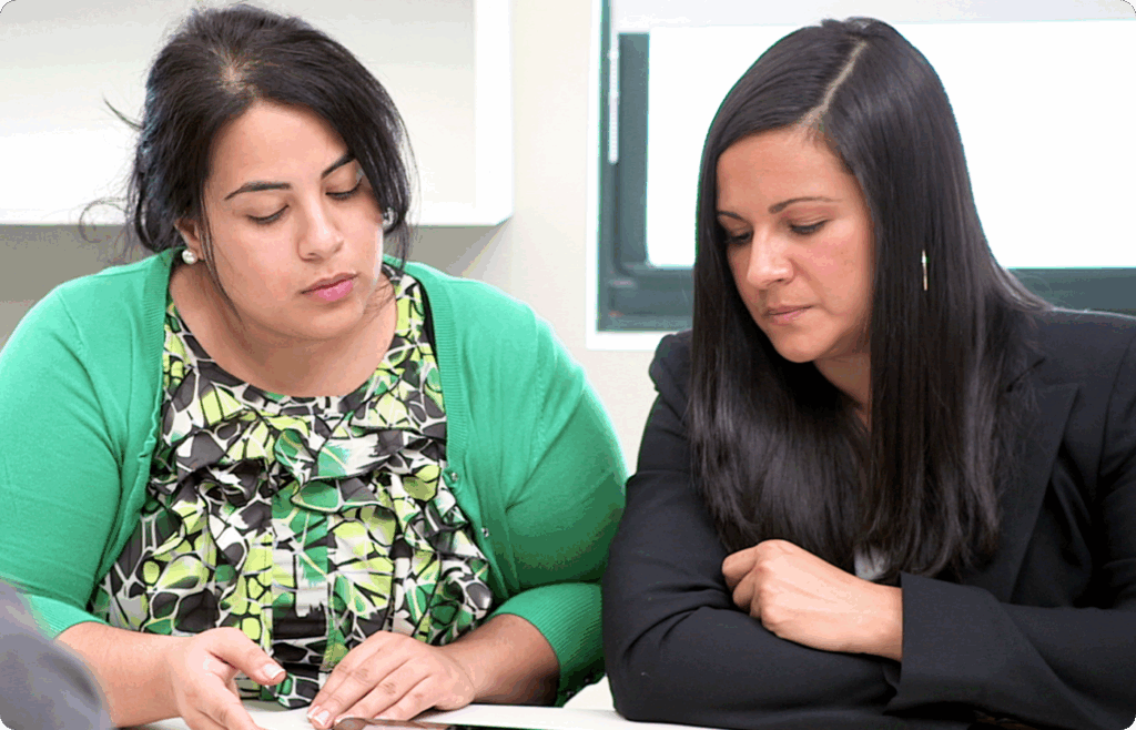 Two women sitting at a table looking down at documents, appearing focused and engaged in discussion or review.