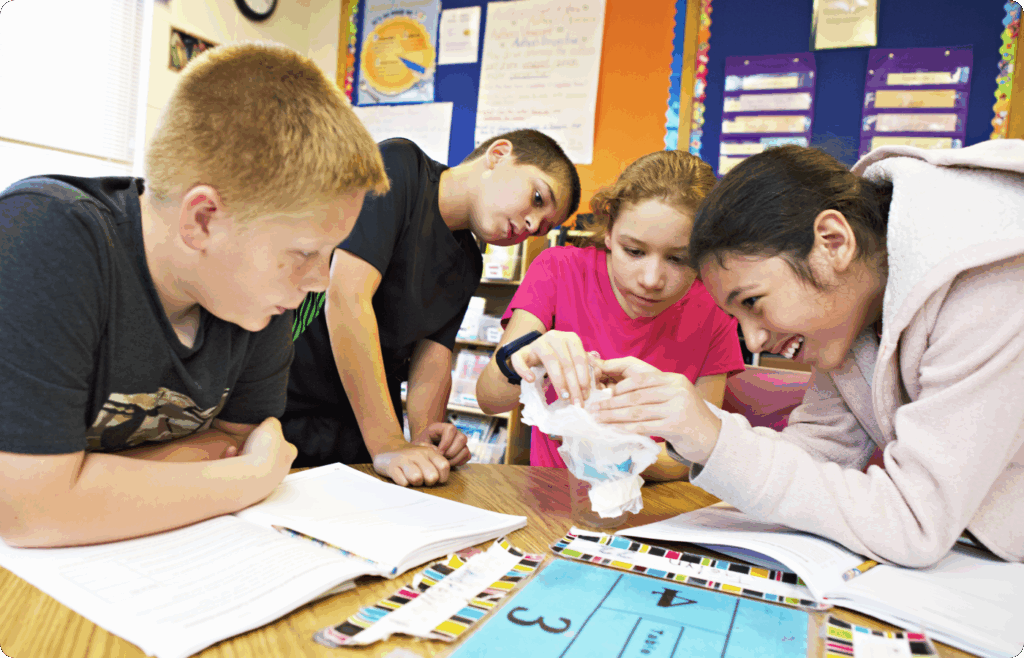 Four children sit at a classroom table examining a small object together, surrounded by open notebooks and colorful classroom decorations.