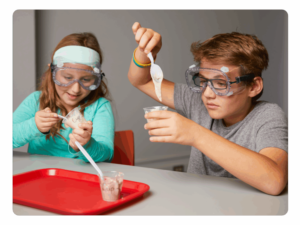 Two children wearing safety goggles conduct an NGSS-inspired science experiment at a table, mixing substances in clear plastic cups with plastic spoons.