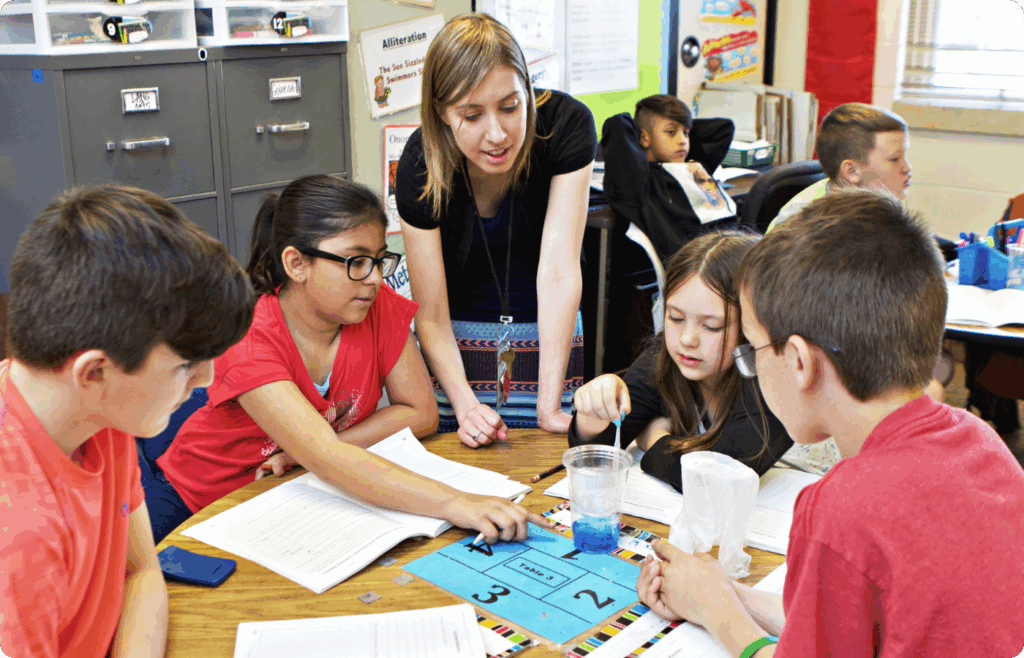 A teacher observes and assists a group of four students working on a science activity at a classroom table.