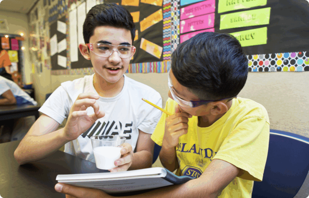 Two boys wearing safety glasses sit at a classroom table. One holds a cup with a white liquid; the other holds a pencil and notebook. Bulletin boards with colorful labels are in the background.