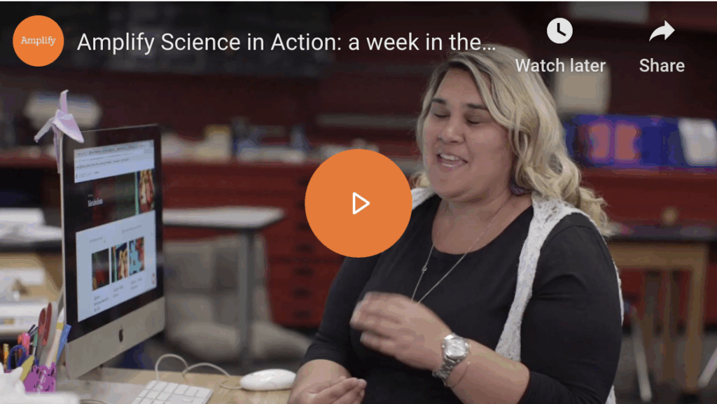A woman smiling and gesturing while speaking, seated in a classroom with computers and educational materials around her, with a paused video screen featuring text about "amplify science.