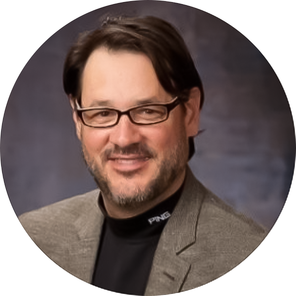 Man with glasses and dark hair wearing a gray blazer and black shirt, posing in front of a dark, blurred background.