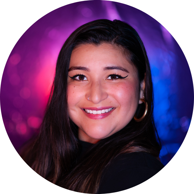A woman with long dark hair, wearing a black top and hoop earrings, smiles in front of a blurred purple and blue background.