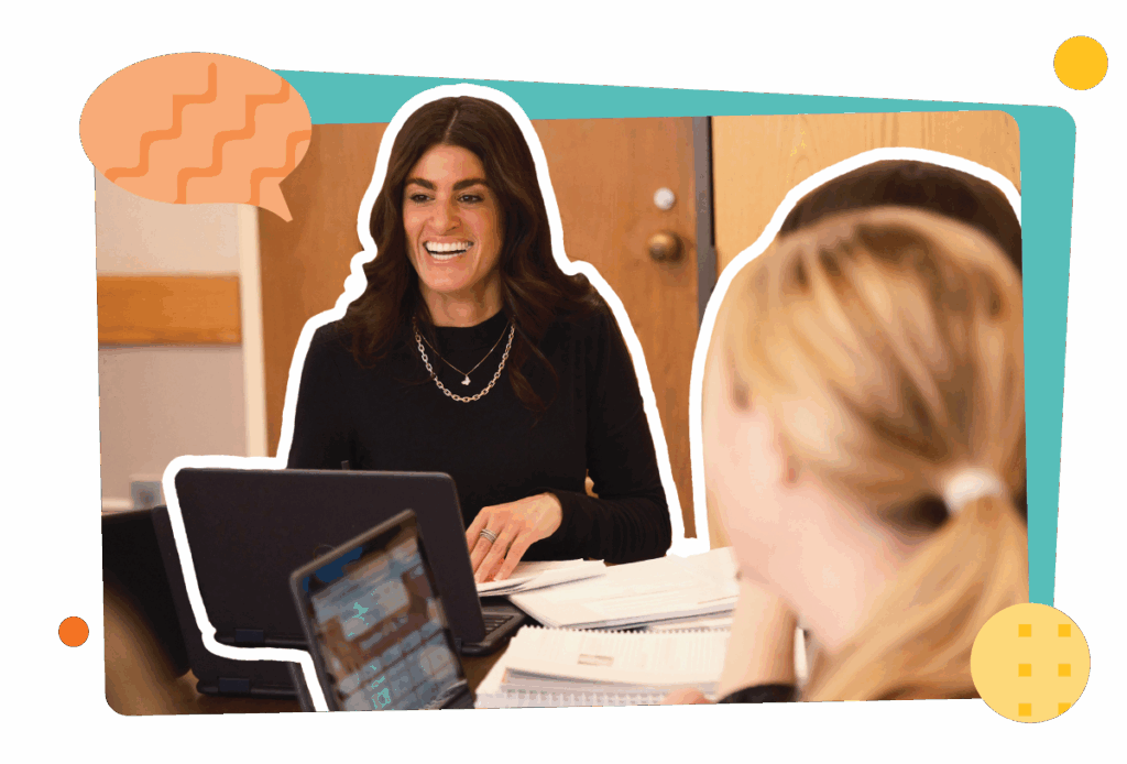 Two women are seated at a table with laptops and papers, engaging in a discussion about program implementation in a classroom or meeting setting.