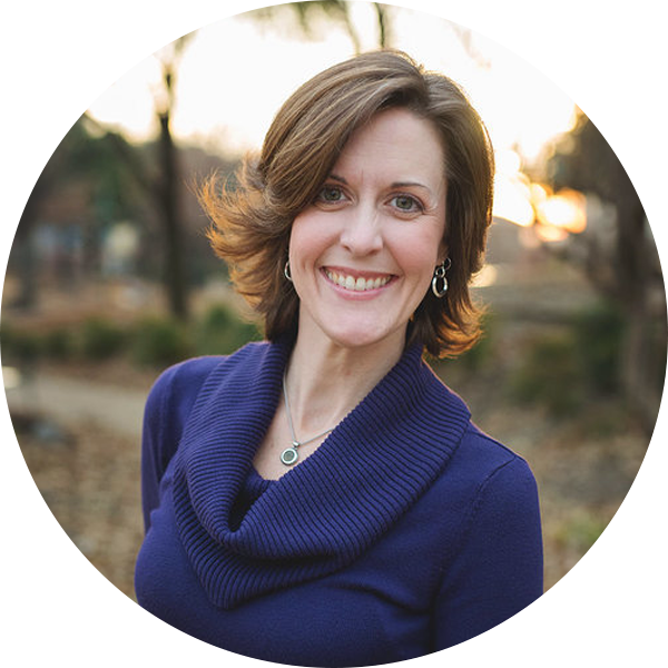 A woman with short brown hair, wearing a purple sweater and silver jewelry, smiles outdoors with a blurred natural background at sunset.