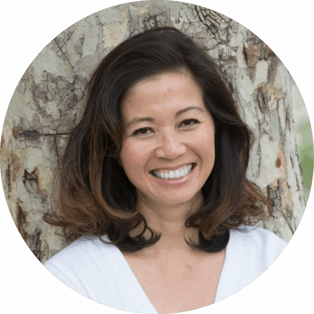 Smiling woman with shoulder-length brown hair stands in front of a tree, wearing a white top.