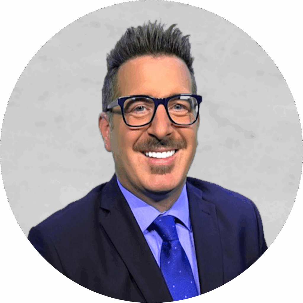 A smiling man with glasses and a dark blue suit with a blue tie, set against a grey background.