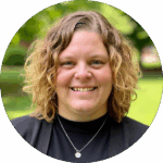 A person with wavy, shoulder-length blonde hair smiles at the camera outdoors, wearing a black top, black jacket, and a round pendant necklace. Greenery is blurred in the background, hinting at a relaxed moment away from studying the science of reading.