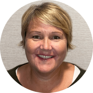 Smiling woman with short blonde hair wearing a dark top, posed against a light-colored, textured background.