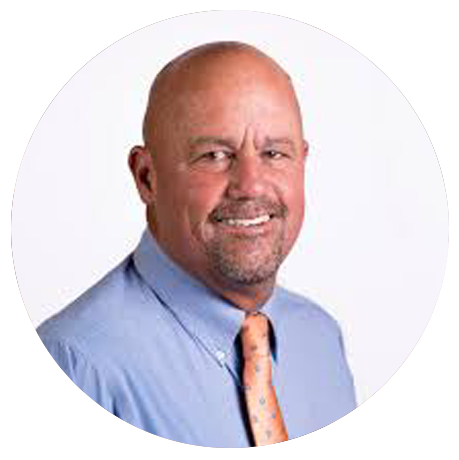 Smiling middle-aged man with a bald head and goatee, wearing a light blue dress shirt and orange patterned tie, posed against a plain white background.