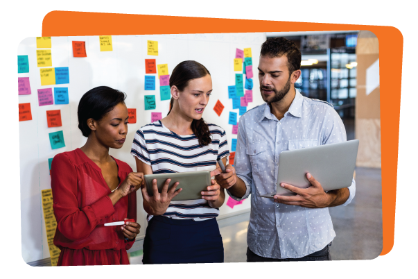 Three people stand in front of a whiteboard with colorful sticky notes, engaging in professional development for teachers as they discuss ideas and collaborate using a tablet and a laptop.