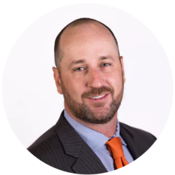 Man with a trimmed beard and shaved head, wearing a dark suit, blue shirt, and orange tie, posing against a plain white background.