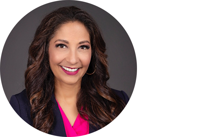 A woman with long, wavy brown hair, wearing a dark blazer and a bright pink blouse, smiles at the camera against a plain gray background.