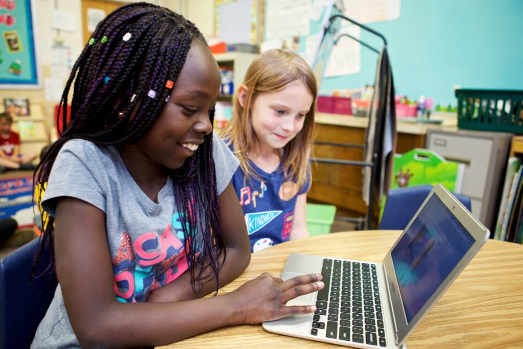 Two girls sit at a classroom table, looking at a laptop screen together. One is typing while the other watches, surrounded by colorful decorations and High Quality Instructional Materials in the background.