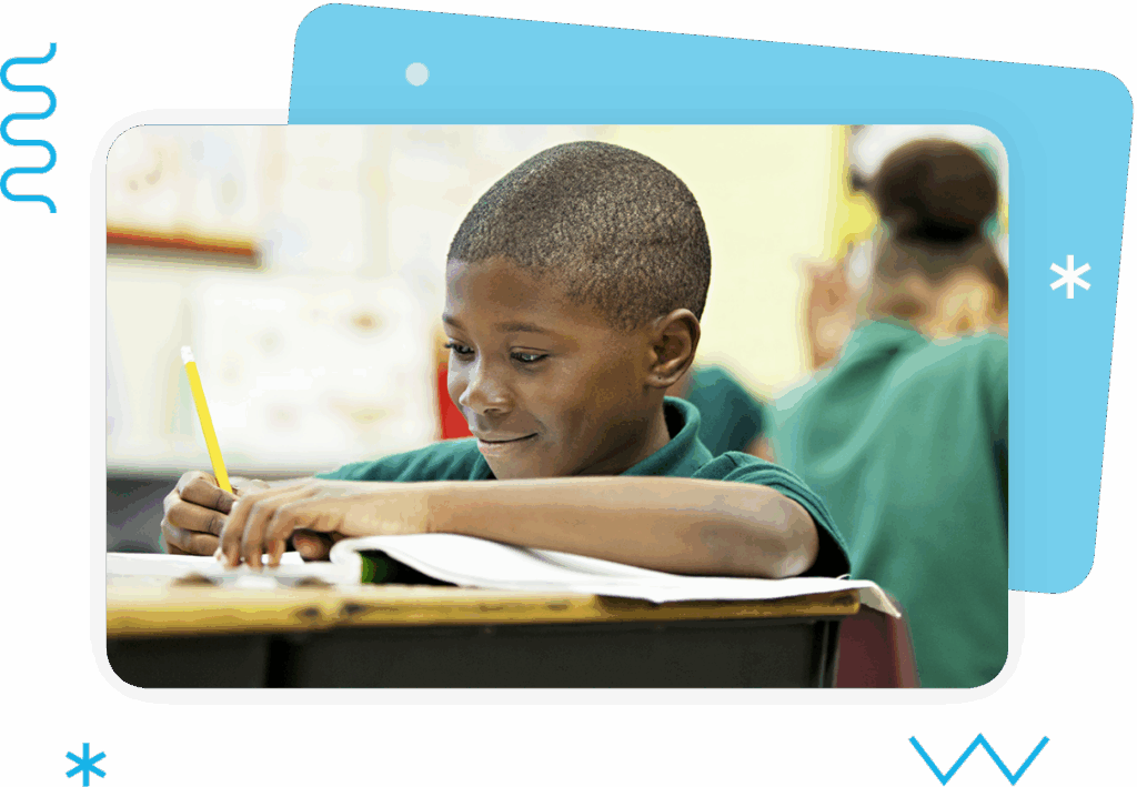 A young boy sits at a desk in a classroom, writing in a notebook with a pencil, while other students are visible in the background.