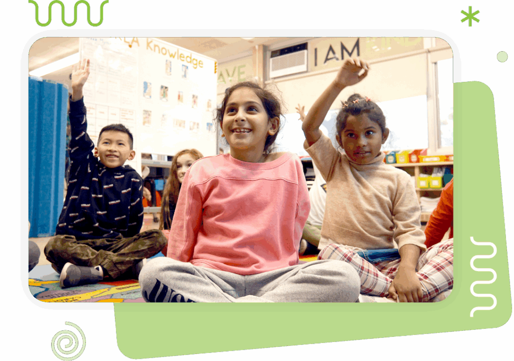 Four children sit cross-legged on a classroom rug, two with hands raised. They appear engaged and attentive, with posters and shelves visible in the background.