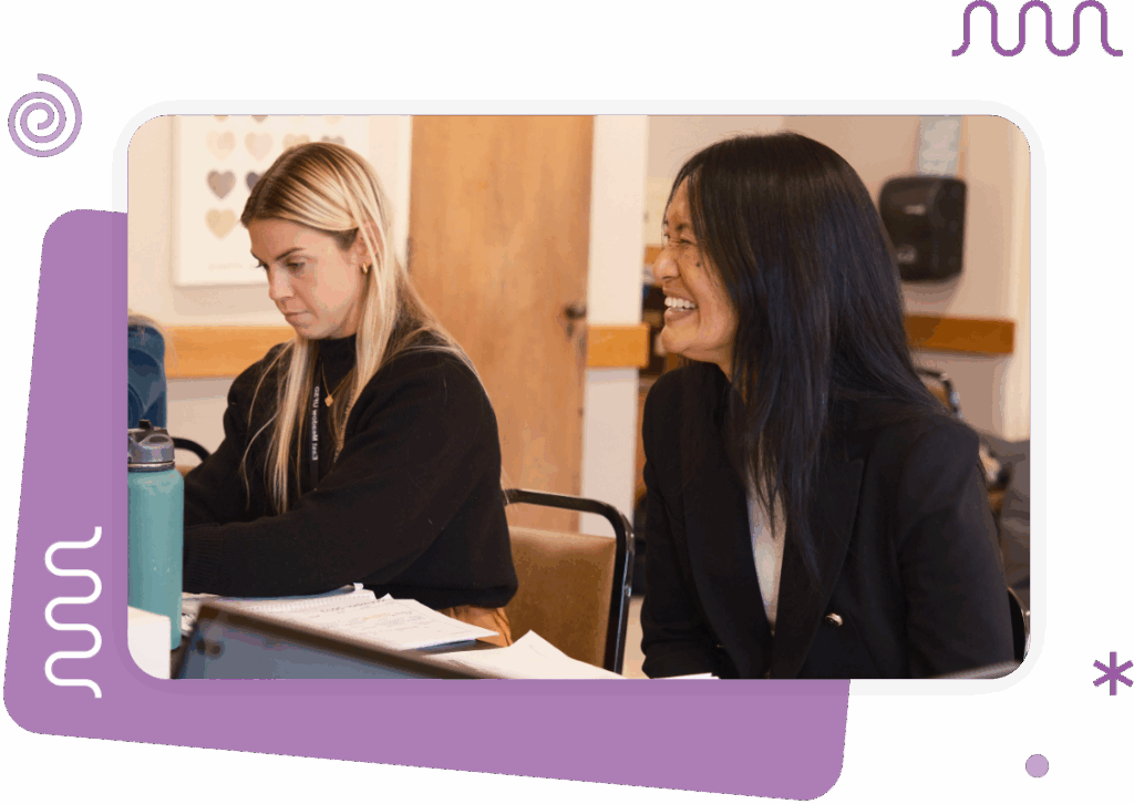 Two women sit indoors at a table; one is focused on her work while the other is smiling. Papers and a water bottle are visible in the foreground.