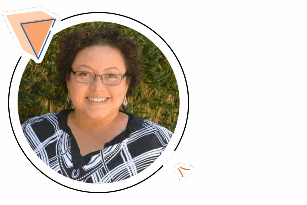 A person with short curly hair and glasses smiles at the camera, wearing a black and white patterned top, with greenery in the background.