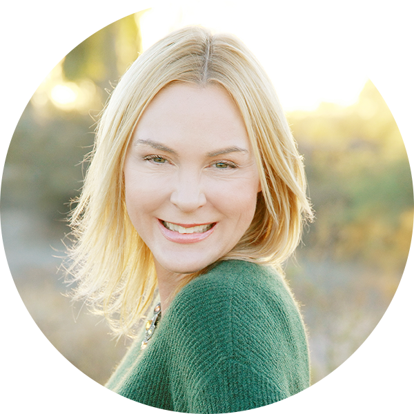 A woman with straight blonde hair wearing a green sweater stands outdoors, smiling at the camera with a blurred natural background.