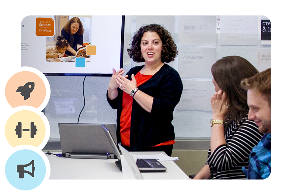 A woman stands and presents to two seated colleagues in an office meeting room, with a laptop and presentation screen visible.