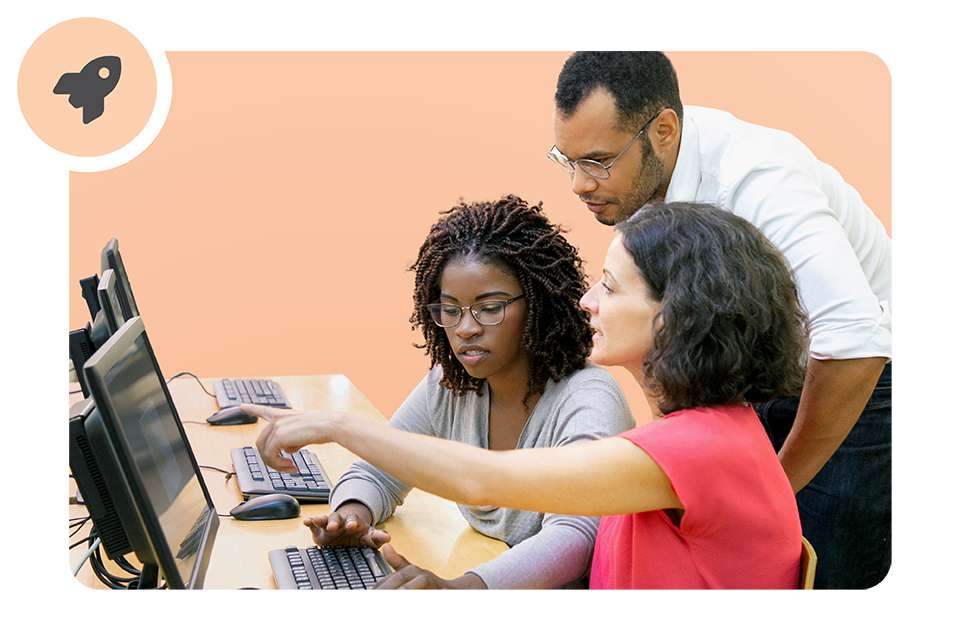 Three people work together at a computer in a classroom; one woman points at the monitor while the others watch attentively.